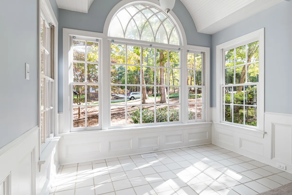 Residential sunroom with a large arched window wall and grid-pattern windows overlooking a wooded backyard